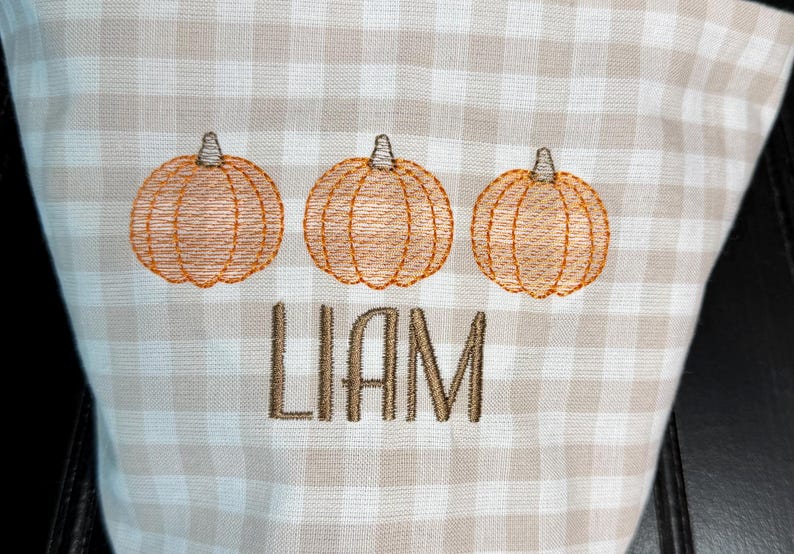 May include: A beige and white checkered fabric with three embroidered orange pumpkins and the name "LIAM" in brown thread. The pumpkins are detailed with stitched lines and stems.