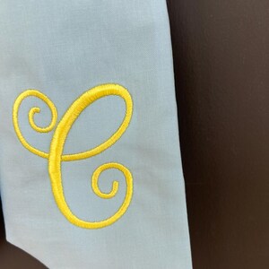 May include: Light blue fabric with a yellow embroidered letter "C". The letter is in a cursive style. The fabric is a solid color and appears to be a smooth material. The background is a dark brown color.