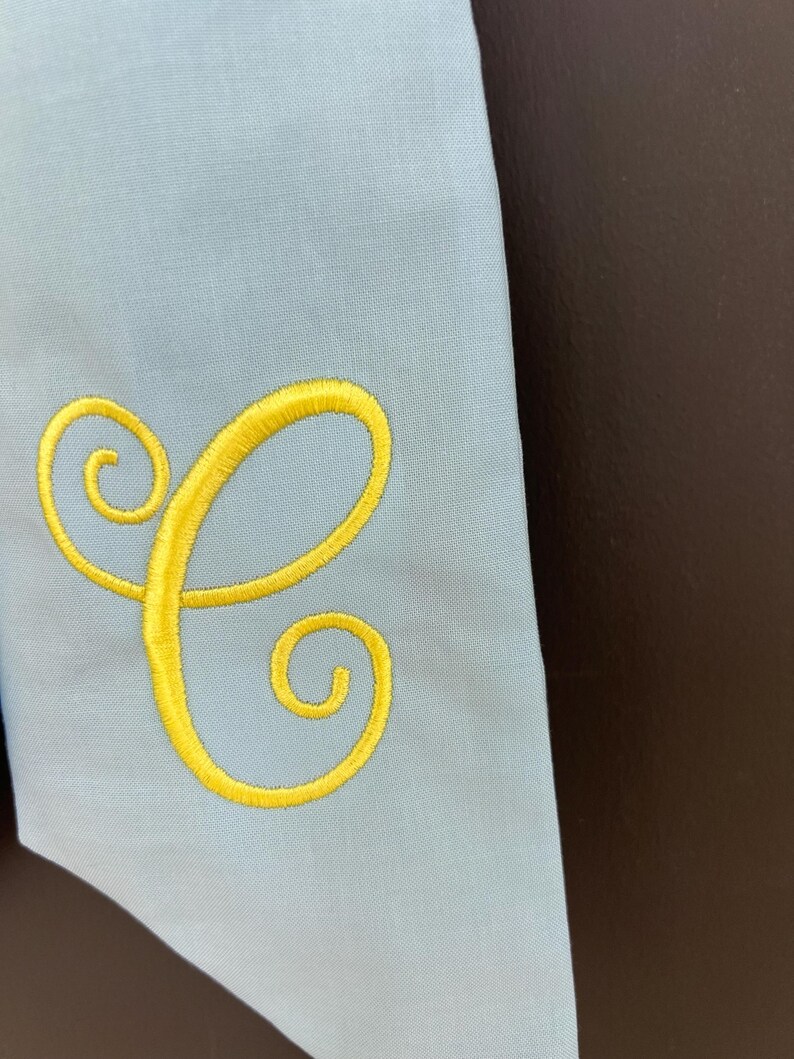 May include: Light blue fabric with a yellow embroidered letter "C". The letter is in a cursive style. The fabric is a solid color and appears to be a smooth material. The background is a dark brown color.