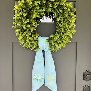 May include: A round, green eucalyptus wreath with a light blue ribbon. The ribbon is embroidered with a white flower and the yellow letter "C". The wreath is hanging on a gray door.