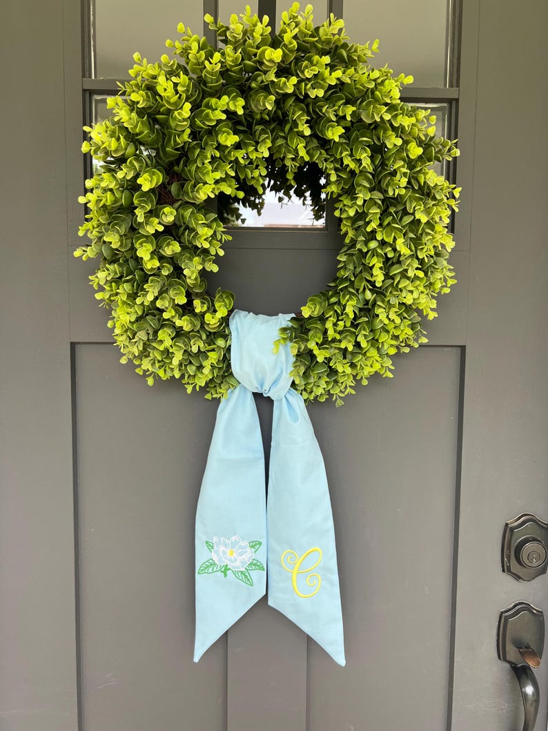 May include: A round, green eucalyptus wreath with a light blue ribbon. The ribbon is embroidered with a white flower and the yellow letter "C". The wreath is hanging on a gray door.