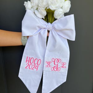 May include: A bouquet of white flowers tied with a large white bow. The bow has two long tails with pink embroidered text that reads "HOCO 2024" and a monogram. The flowers are white and the background is dark gray.