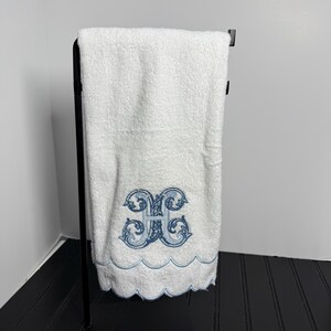 May include: White hand towel with a blue embroidered monogram and scalloped edge, hanging on a black metal towel rack. The monogram is an ornate, stylized letter "H".