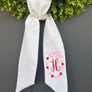 May include: White fabric ribbon with a pink bow and a red embroidered letter H surrounded by pink, red, and gold hearts.