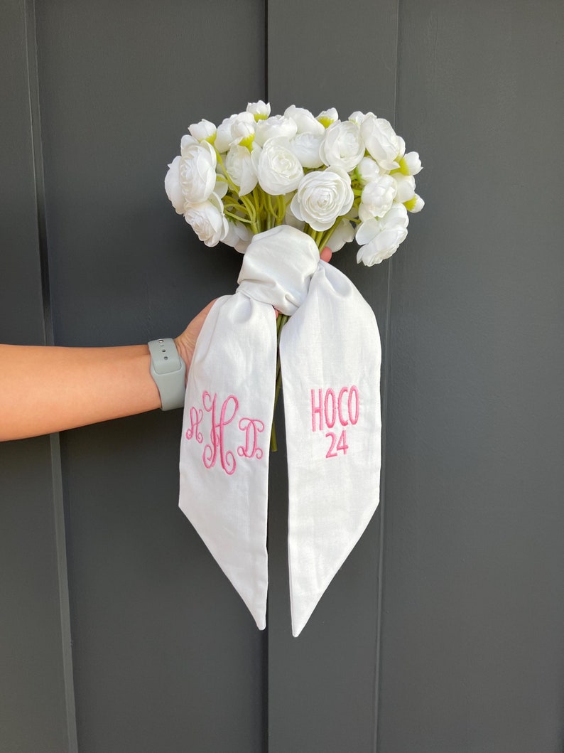 May include: A bouquet of white flowers tied with a white ribbon. The ribbon has pink embroidered text that reads "K D" and "HOCO 24". The flowers are clustered together, creating a full, round shape.