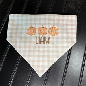 May include: A tan and white checkered dog bandana with three embroidered orange pumpkins and the name "LIAM" in brown thread. The bandana is triangular in shape and made of fabric.