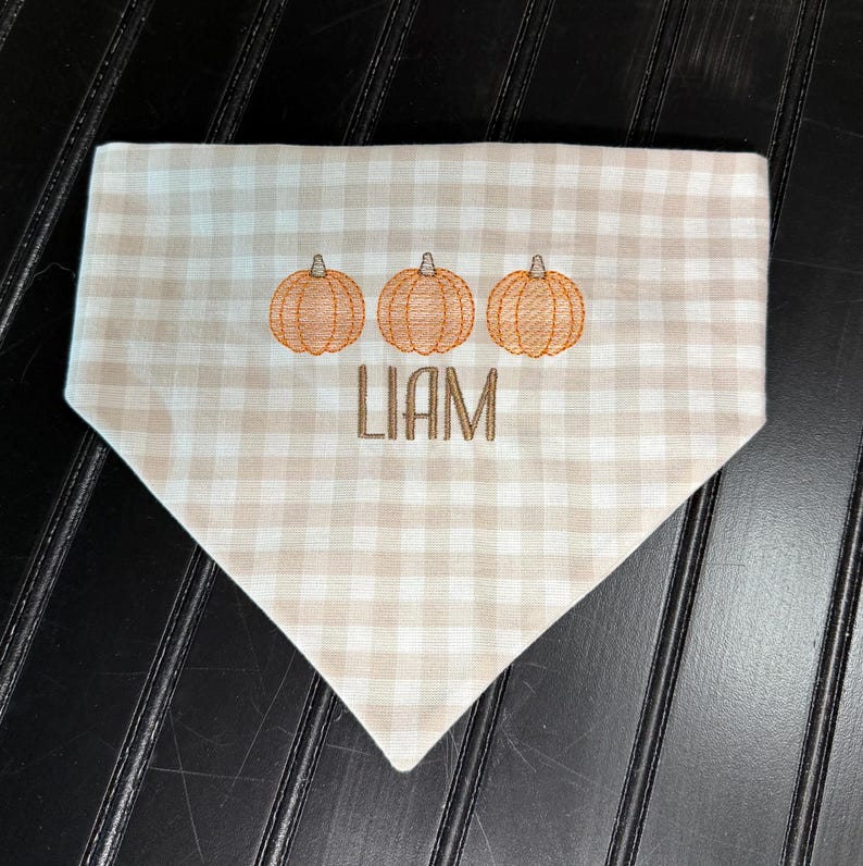 May include: A tan and white checkered dog bandana with three embroidered orange pumpkins and the name "LIAM" in brown thread. The bandana is triangular in shape and made of fabric.