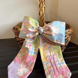K&ouml;nnte beinhalten: Eine dekorative Osterkorb-Schleife mit einem Patchwork-Design mit Hasen, Karotten und floralen Mustern. Die Schleife ist an einem Weidenkorb befestigt. Die Schleife hat eine monogrammierte Initiale in rosa Faden.