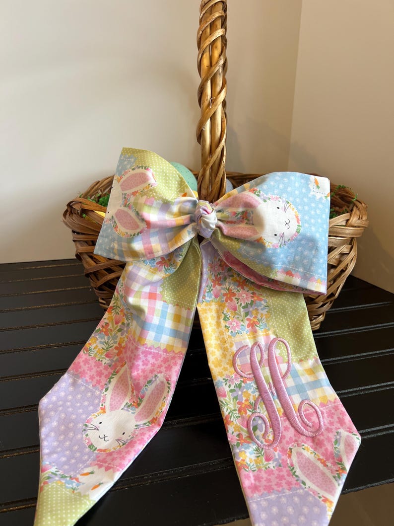 K&ouml;nnte beinhalten: Eine dekorative Osterkorb-Schleife mit einem Patchwork-Design mit Hasen, Karotten und floralen Mustern. Die Schleife ist an einem Weidenkorb befestigt. Die Schleife hat eine monogrammierte Initiale in rosa Faden.