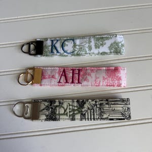 May include: Three fabric keychains with embroidered initials. The top keychain features a white and green floral pattern with the initials "KC" in blue. The middle keychain has a pink pattern with the initials "AH" in purple. The bottom keychain has a black and white pattern with the initials "PD" in green.