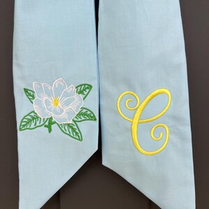 May include: Two light blue fabric sashes. One sash features a white and light blue embroidered magnolia flower with green leaves. The other sash has a yellow embroidered letter "C".