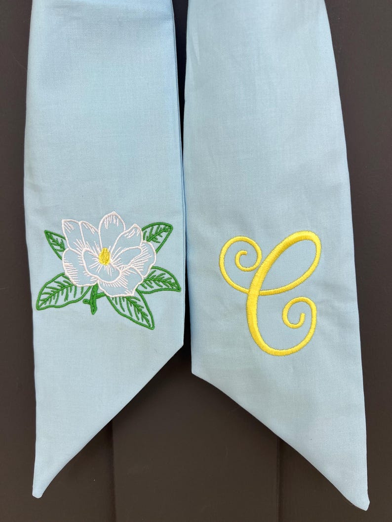 May include: Two light blue fabric sashes. One sash features a white and light blue embroidered magnolia flower with green leaves. The other sash has a yellow embroidered letter "C".
