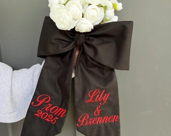 May include: A bouquet of white flowers attached to a large black fabric bow. The bow features red embroidered text on its two long tails, reading Prom 2025 on the left and Lily & Brennen on the right, with a knot center holding the flowers.