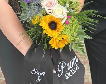May include: A floral bouquet featuring yellow sunflowers, white and pale pink roses, blue thistle, and green foliage. Two black fabric ribbons hang from the stems, embroidered with white text reading Sean & McKenzie and Prom 2025.