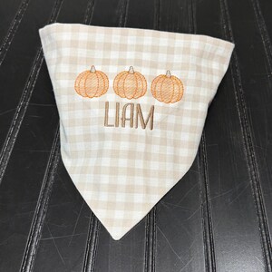 May include: A tan and white checkered bandana with three embroidered orange pumpkins and the name "LIAM" in brown thread. The bandana is folded into a triangle shape.