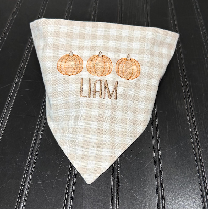 May include: A tan and white checkered bandana with three embroidered orange pumpkins and the name "LIAM" in brown thread. The bandana is folded into a triangle shape.