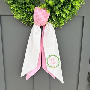 May include: A green wreath with a pink and white gingham bow and white fabric streamers. The streamers have a green floral wreath with a monogram in the centre. The wreath is hanging on a grey door.