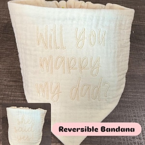 May include: Cream-colored, reversible bandana with embroidered text. One side reads "Will you marry my dad?" and the other side says "She said yes!". The bandana has a soft, textured appearance and a pink label that says "Reversible Bandana".
