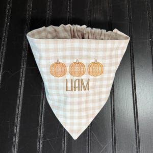 May include: A tan and white gingham patterned pet bandana with three embroidered orange pumpkins and the name "LIAM" in brown thread. The bandana has a gathered top edge.