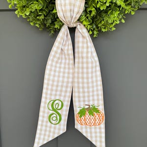May include: A green wreath with a tan and white checkered ribbon. The ribbon has an embroidered green letter "S" and an orange pumpkin with green leaves. The ribbon is tied in a knot.