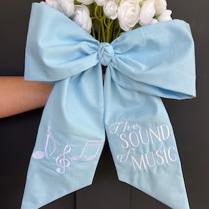 May include: A light blue bow with white embroidered musical notes and the text "The Sound of Music". The bow is tied around a bouquet of white flowers.