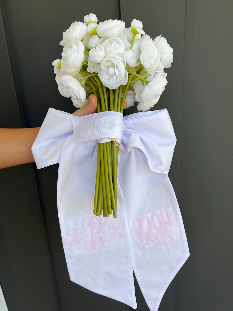 May include: A bouquet of white flowers with green stems, tied with a white ribbon. The ribbon has pink text and a monogram. The flowers are clustered together, creating a full and lush appearance.