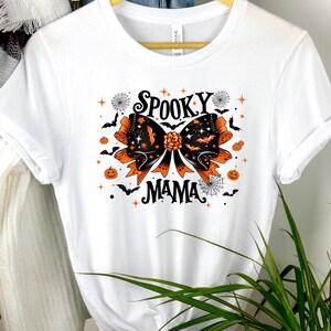 May include: White t-shirt with a Halloween graphic. The design includes a large bow with orange and black details, bats, spiders, and pumpkins. The words "SPOOKY MAMA" are printed in black.