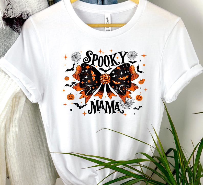 May include: White t-shirt with a Halloween graphic. The design includes a large bow with orange and black details, bats, spiders, and pumpkins. The words "SPOOKY MAMA" are printed in black.