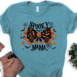 May include: A turquoise t-shirt with a Halloween design. It features a black and orange bow with bats, stars, and the words "Spooky Mama". Orange pumpkins and spiderwebs are also included in the design.