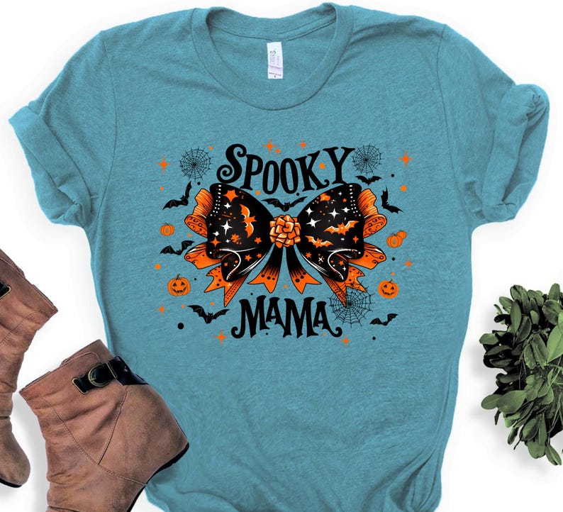 May include: A turquoise t-shirt with a Halloween design. It features a black and orange bow with bats, stars, and the words "Spooky Mama". Orange pumpkins and spiderwebs are also included in the design.