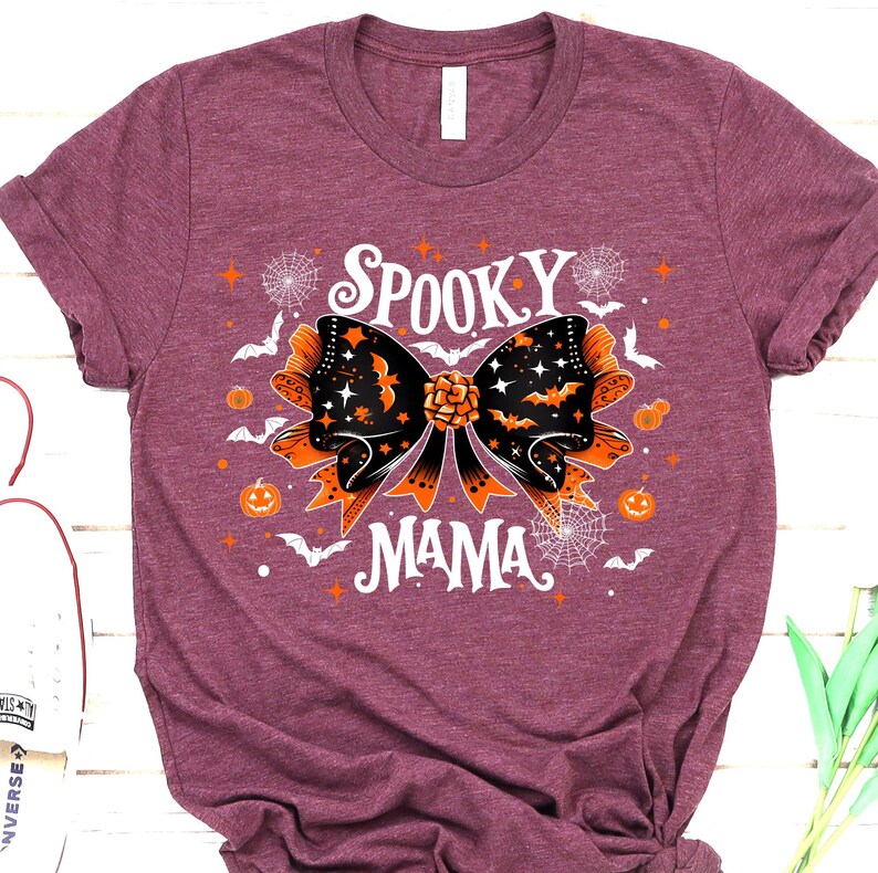 May include: Burgundy t-shirt with a Halloween design. The graphic features a black and orange bow with bats, stars, and the words "Spooky Mama" in white. Additional elements include pumpkins, spiderwebs, and bats.