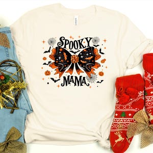 May include: Cream t-shirt with a Halloween graphic. The design features a large orange and black bow with bats, stars, and the words "Spooky Mama". Additional Halloween elements include pumpkins and spiderwebs.