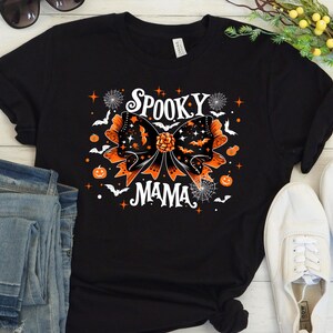 May include: Black t-shirt with a Halloween design. The graphic includes an orange and black bow with bats, stars, and the words "Spooky Mama" in white. Pumpkins and spiderwebs complete the Halloween theme.