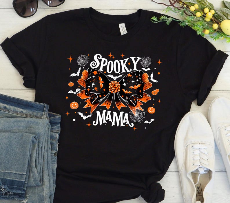 May include: Black t-shirt with a Halloween design. The graphic includes an orange and black bow with bats, stars, and the words "Spooky Mama" in white. Pumpkins and spiderwebs complete the Halloween theme.