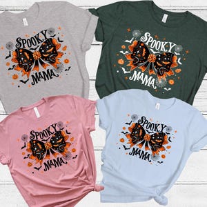 May include: Four t-shirts in grey, green, pink, and light blue. Each shirt displays a Halloween graphic with a black bow, bats, pumpkins, and the words "Spooky Mama".