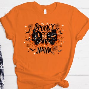 May include: Orange t-shirt with a black and orange graphic. The graphic features a large bow with bats, stars, and the words "Spooky Mama". Other Halloween-themed elements include spiderwebs and pumpkins.
