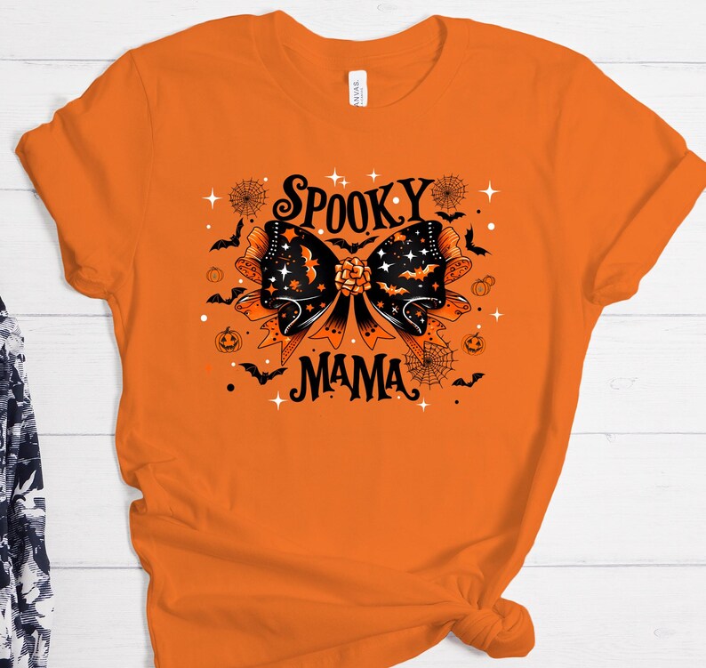 May include: Orange t-shirt with a black and orange graphic. The graphic features a large bow with bats, stars, and the words "Spooky Mama". Other Halloween-themed elements include spiderwebs and pumpkins.