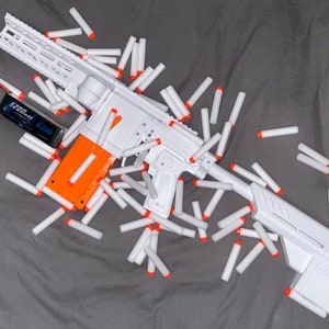 May include: A white and orange toy gun with a black battery pack and a large number of white foam darts scattered around it.