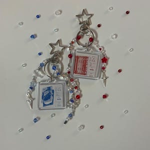 May include: Two keychains with star-shaped charms and red, white and blue beads. Each keychain features a clear square charm with a printed image inside. One charm has a red image of a building, and the other charm has a blue image of a building.