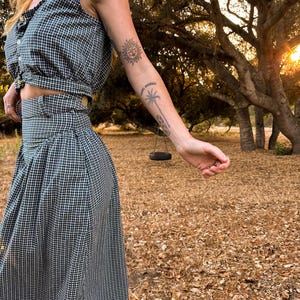 May include: A woman wearing a black and white gingham crop top and matching skirt. She is standing in a field with trees in the background. The woman has a sun and palm tree tattoo on her arm.