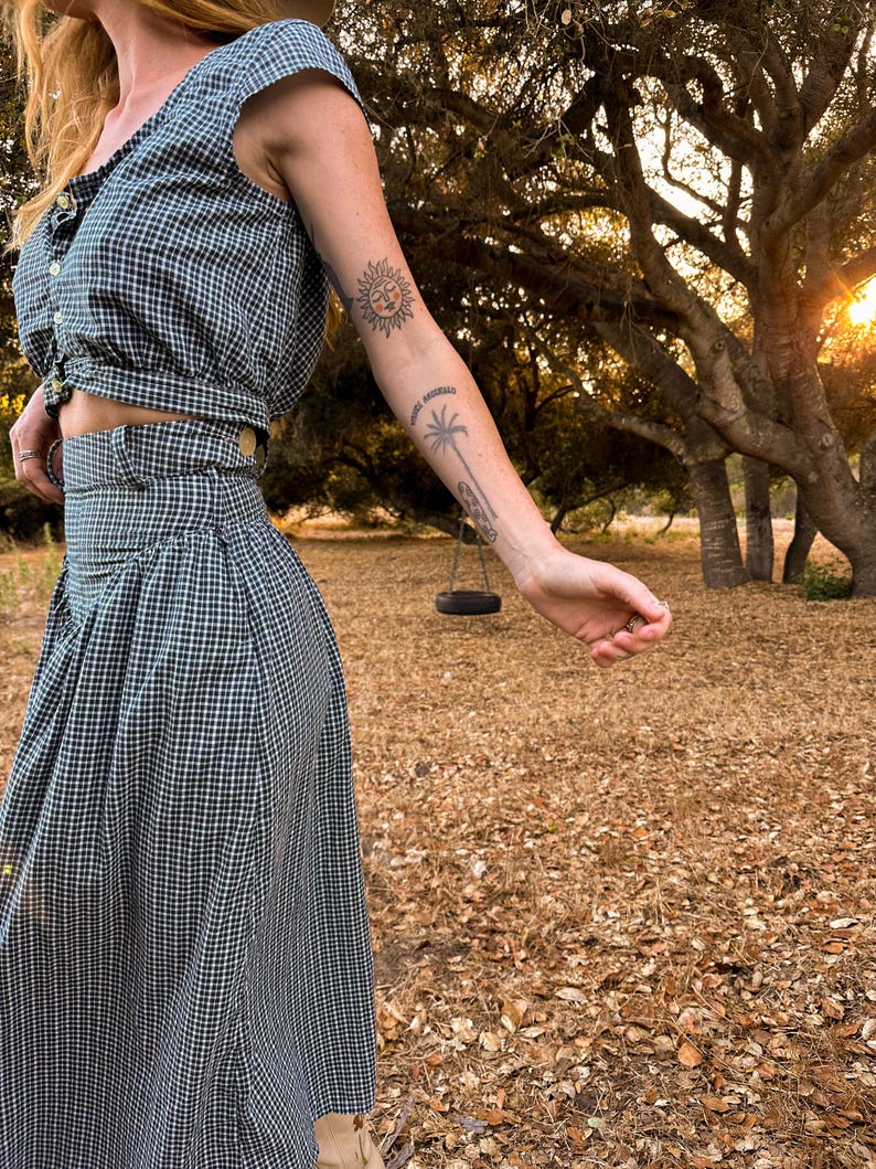 May include: A woman wearing a black and white gingham crop top and matching skirt. She is standing in a field with trees in the background. The woman has a sun and palm tree tattoo on her arm.