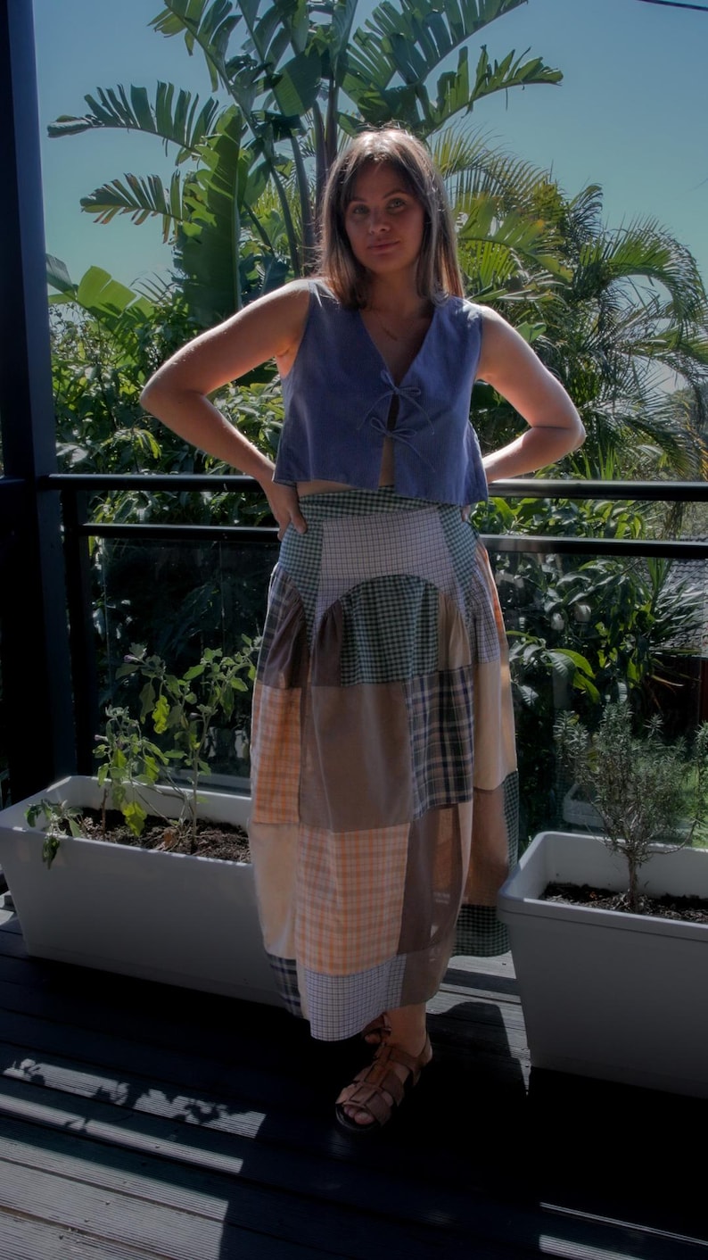 May include: A woman wearing a light blue sleeveless top with a tie at the neckline and a patchwork skirt with brown, green, and orange plaid patterns. She is standing on a wooden deck with potted plants in the background.
