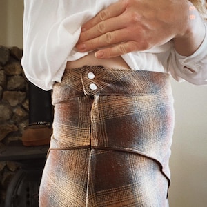 May include: A brown and tan plaid skirt with a brown leather waistband and two white buttons. The skirt is made of a soft, textured fabric.