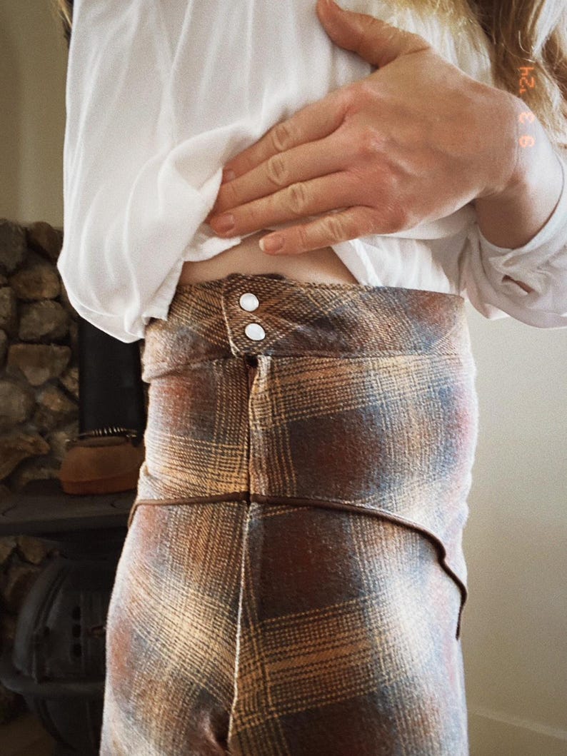 May include: A brown and tan plaid skirt with a brown leather waistband and two white buttons. The skirt is made of a soft, textured fabric.
