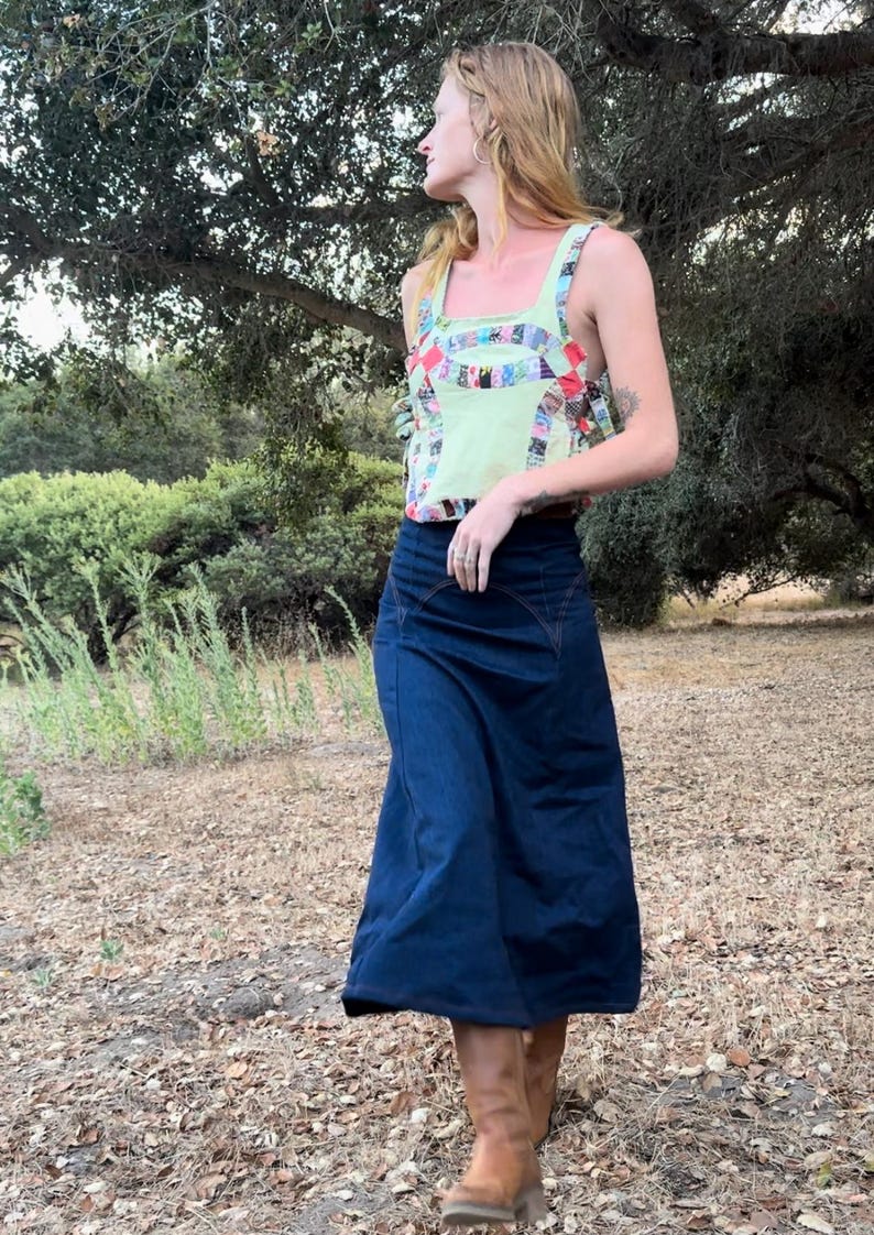 May include: A woman wearing a green and multi-colored patchwork tank top and a dark blue denim skirt. She is standing in a grassy area with trees in the background.
