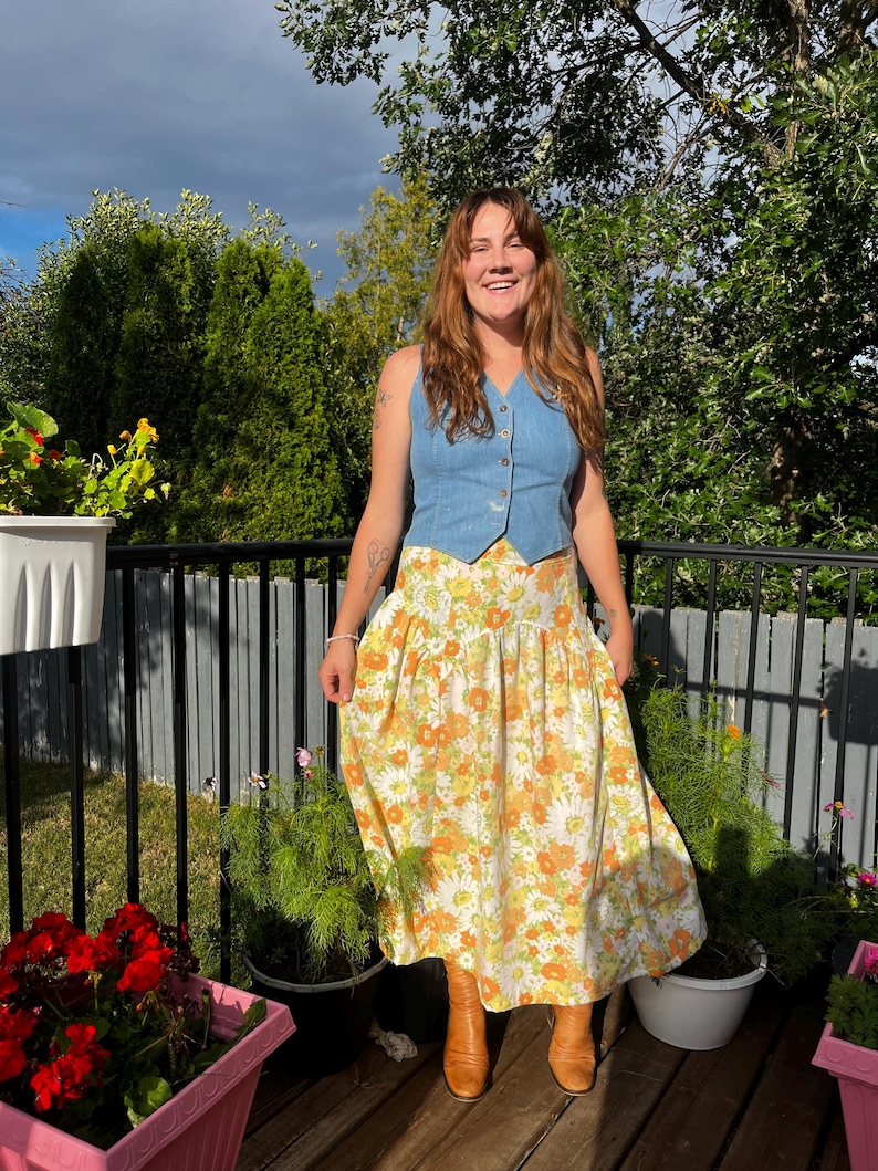 May include: A woman wearing a light blue denim vest over a white and orange floral maxi skirt. She is standing on a wooden deck with potted plants in the background.