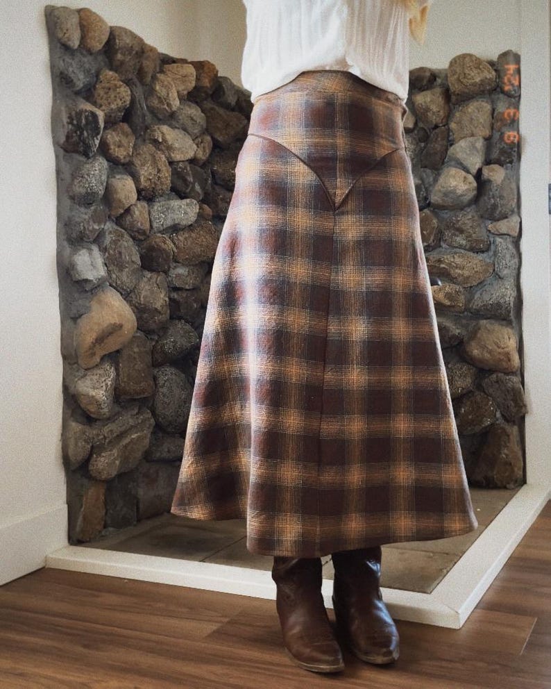May include: A brown and tan plaid skirt with a high waist and a slight A-line shape. The skirt is made of a soft, woven fabric and has a brown leather trim.