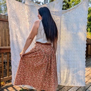 May include: A woman wearing a white sleeveless top and a brown floral midi skirt with a brown belt. She is wearing brown cowboy boots and has long black hair. She is standing in front of a white lace curtain.