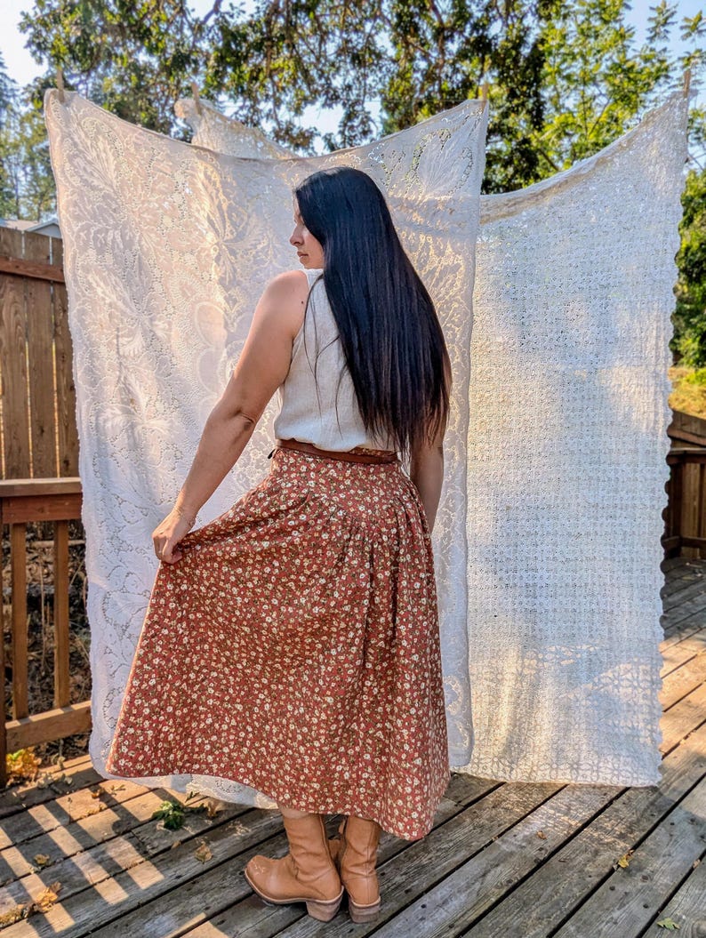 May include: A woman wearing a white sleeveless top and a brown floral midi skirt with a brown belt. She is wearing brown cowboy boots and has long black hair. She is standing in front of a white lace curtain.