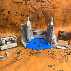 Op de afbeelding: Een bordspelscène met een miniatuurmodel van de Argonath-beelden, een blauwe waterpartij en verschillende speelkaarten. De scène speelt zich af op een kaart met tekst. Drie kleine boten liggen in het water.